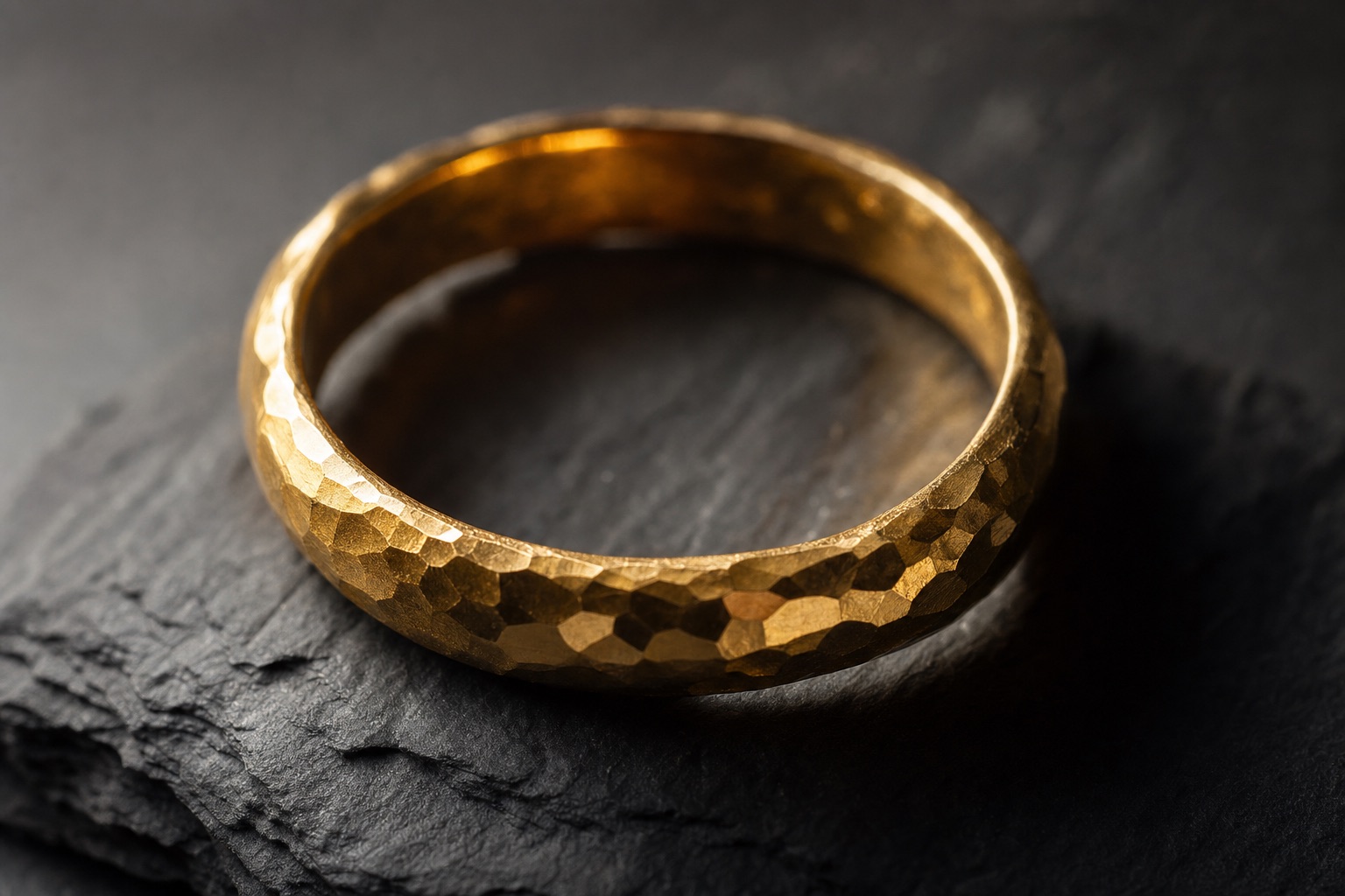 Macro view of a hand-hammered 22-karat gold band on charcoal slate, facets catching warm light
